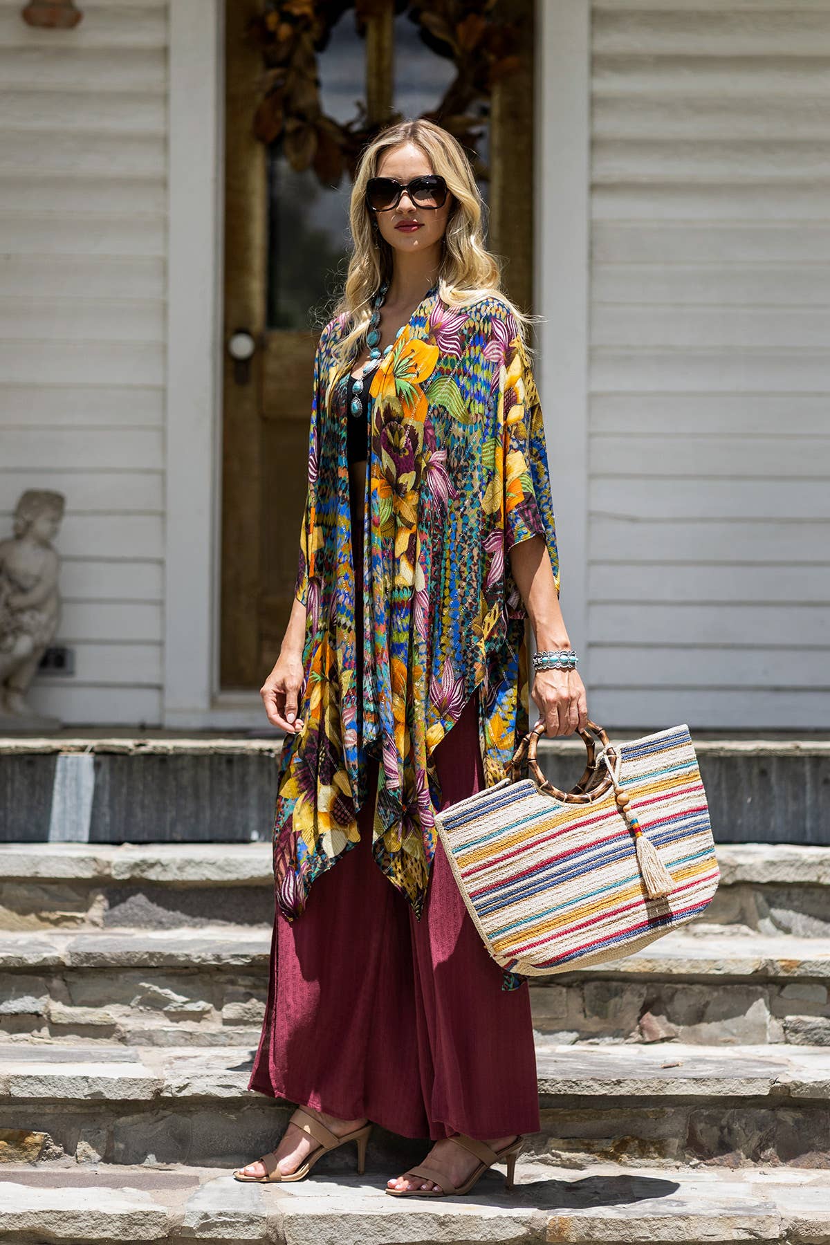 Woman standing on stone steps wearing a colorful floral kimono, maroon wide-leg pants, sunglasses, and holding a striped.