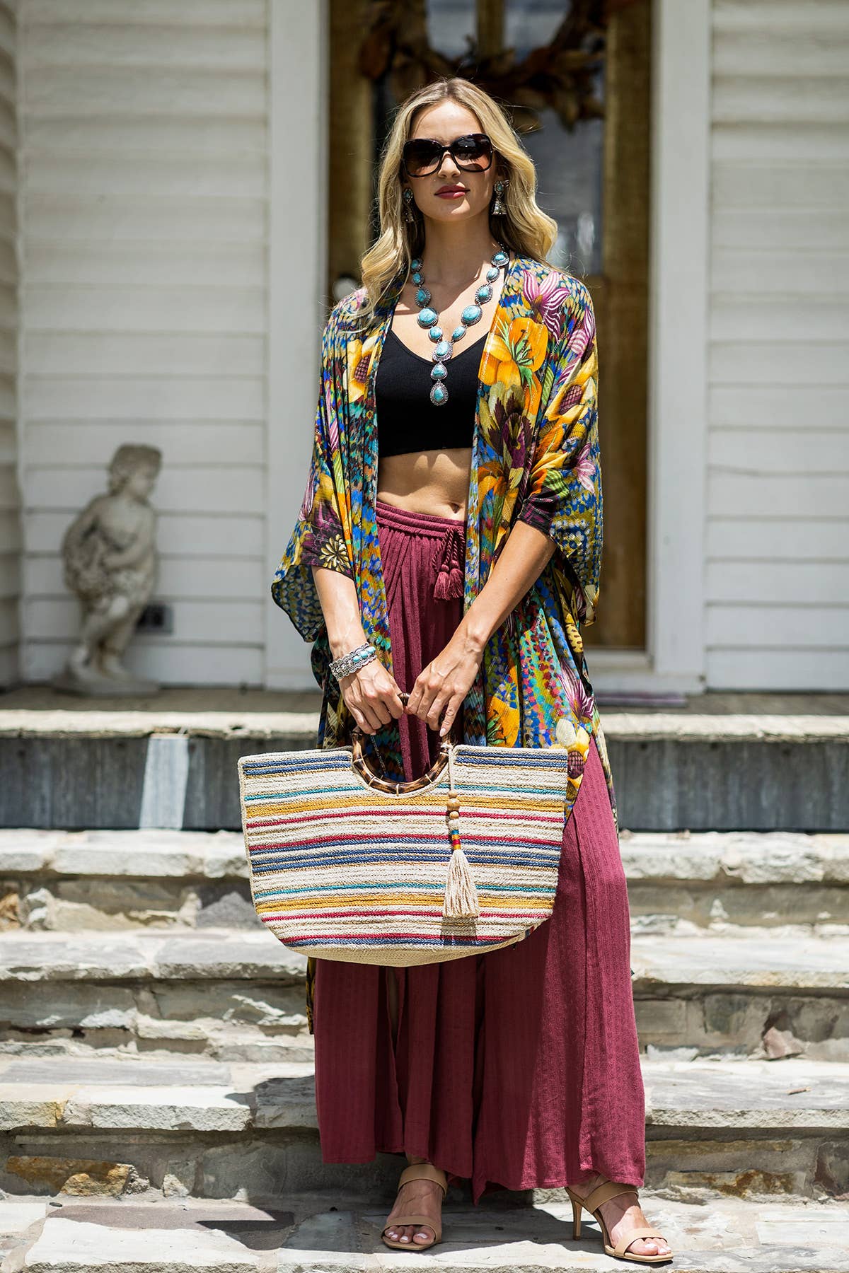 Woman wearing sunglasses, a floral kimono, black crop top, maroon skirt, holding a striped bamboo and jute bag with round.