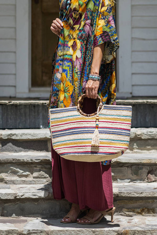 Hand holding a large bamboo jute bag with multicolored horizontal stripes and round bamboo handles, tassel detail hanging.