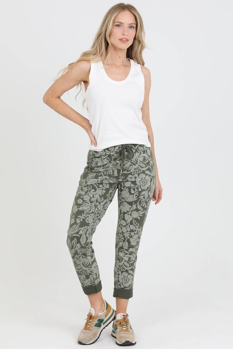 Olive green jogger pants with dark floral print, cuffed ankles, worn with white tank and beige sneakers.