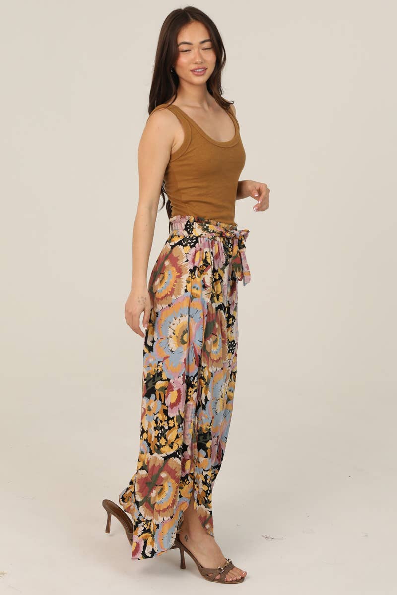 Woman facing side wearing a brown tank top and Angie colorful floral pants on a beige background swhwing the length with heels