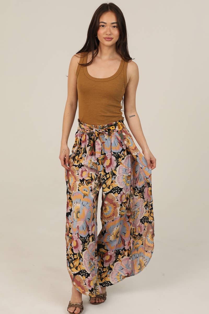 Woman facing forward wearing a brown tank top and Angie colorful floral pants on a beige background showing the width of the pants
