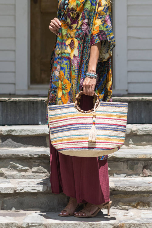 Striped bamboo and jute tote bag with round wooden handles and white tassel, held by person wearing a colorful patterned.